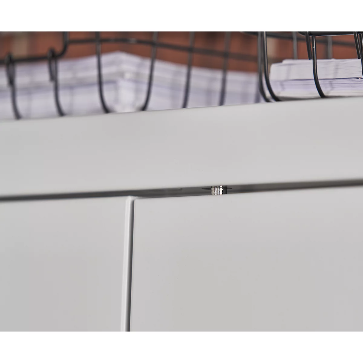 Close-up of white cabinet doors, a narrow silver metal trim and a wire basket with papers in the background.