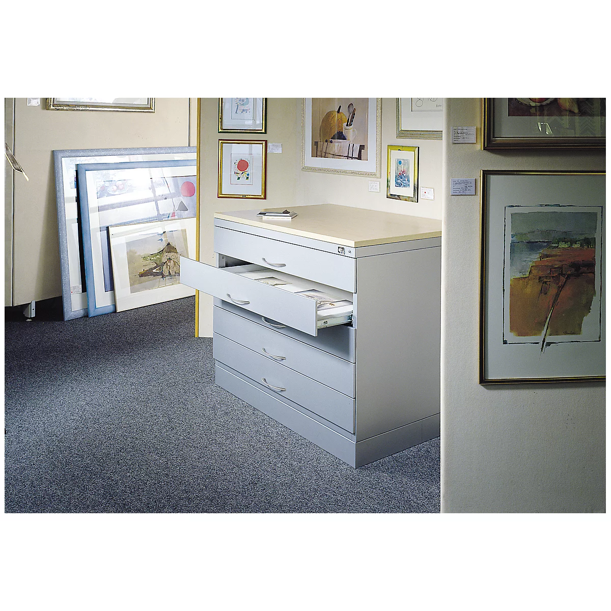 Gray drawer cabinet, one drawer open. Artwork on walls. Gray carpeted floor.