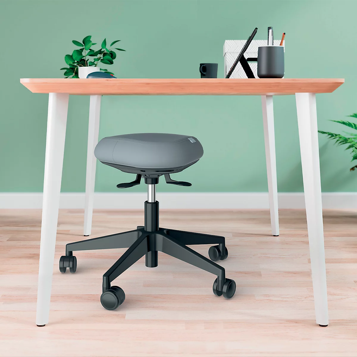 Schreibtisch mit grauen Bürostuhl, auf Rollen. Auf dem Tisch: Topfpflanze, Becher, Stiftehalter und Tablet.