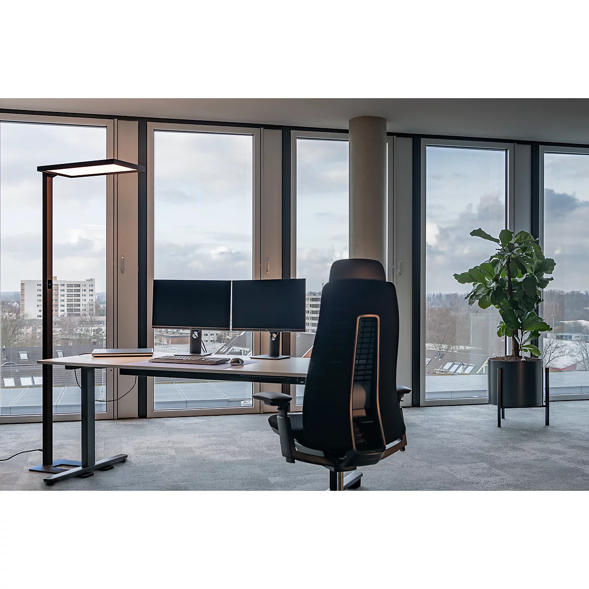 Ein moderner Schreibtisch mit zwei Bildschirmen vor einer Fensterfront. Ein Bürostuhl, Stehlampe und eine Topfpflanze ergänzen das Bild.