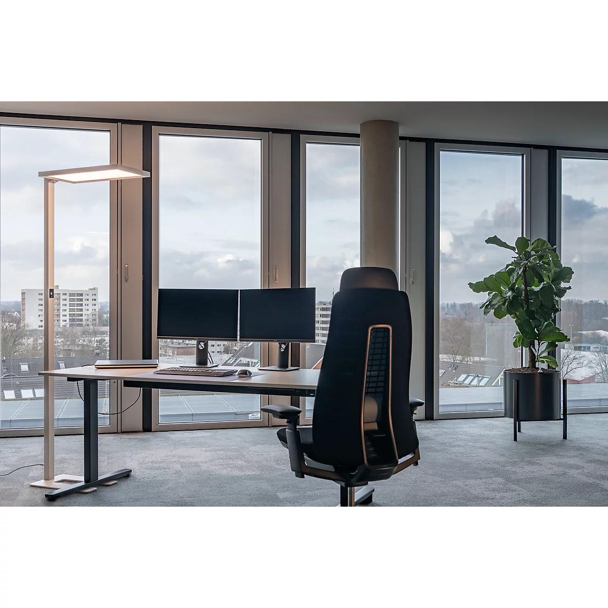 Büro mit Fensterfront, Schreibtisch, Monitoren, Stehleuchte und Bürostuhl mit Blick auf eine Stadt.