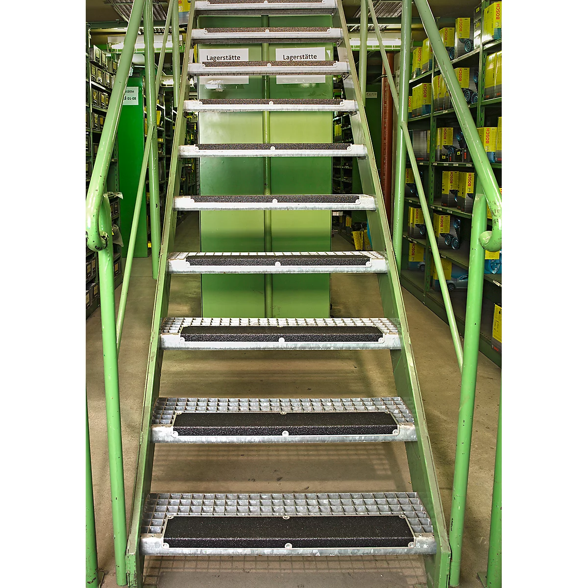Metal staircase in a warehouse, with green railings and black anti-slip strips on the steps. Shelves in the background.