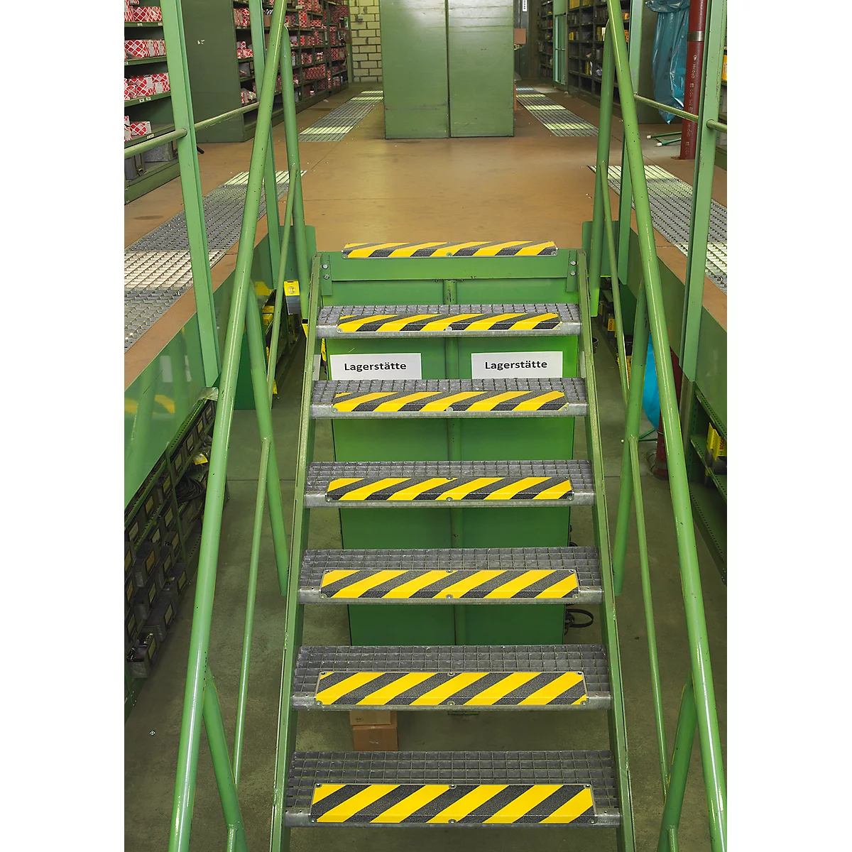 Green metal stairs with yellow and black warning markings in a warehouse. Two signs reading "Lagerstätte." Shelves in the background.