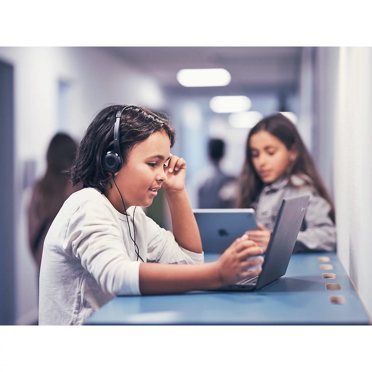 Een kind met een koptelefoon typt op een laptop. Een ander kind werkt op een tablet op de achtergrond.