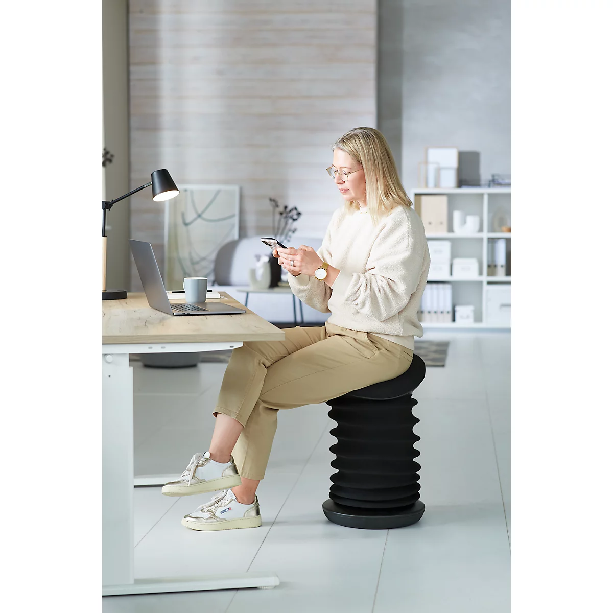 A woman sits at a desk looking at her smartphone. She is wearing beige pants, a cream sweater, and white sneakers. There is a laptop next to her.