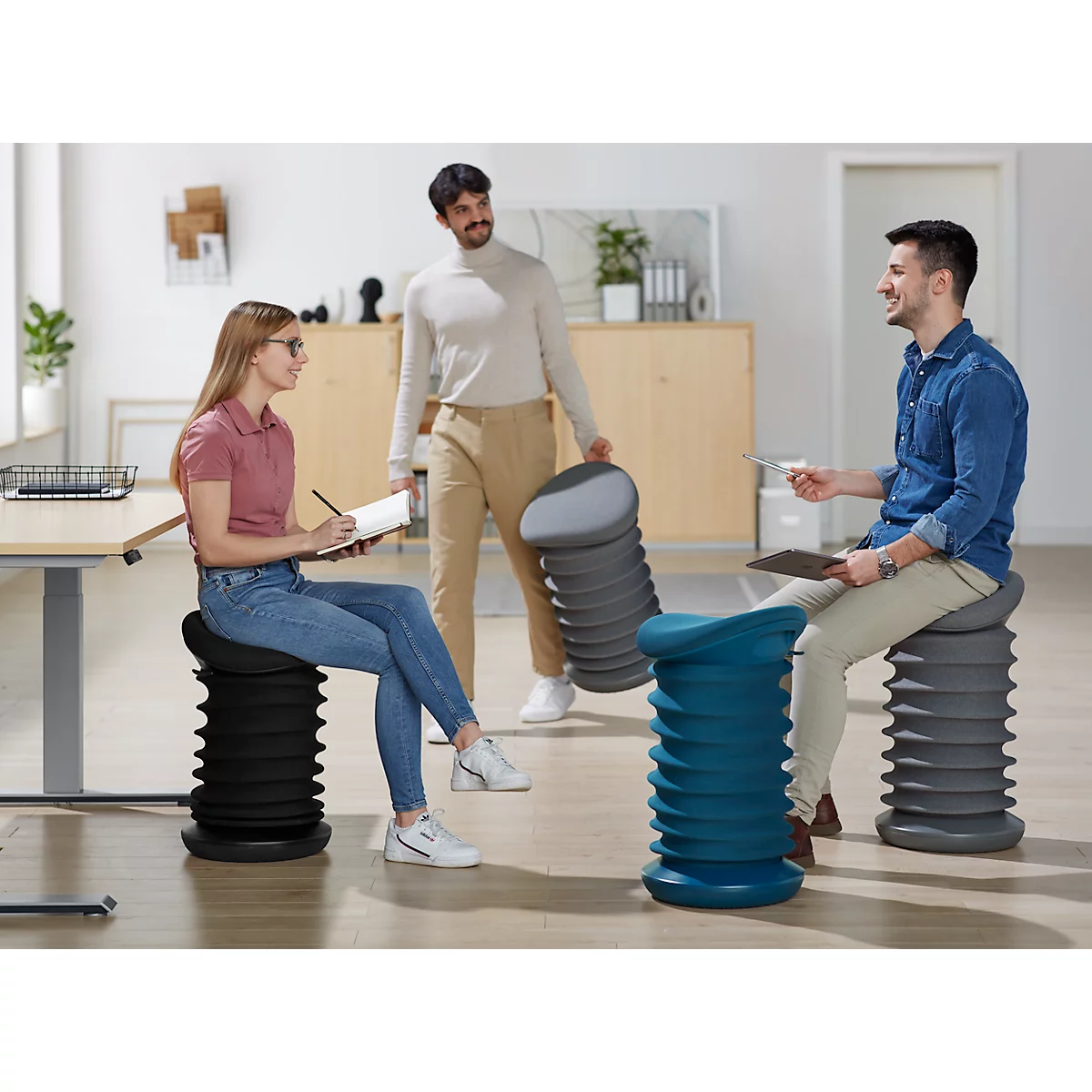 Three people are sitting and standing around standing stools. One person is taking notes, the other is holding a tablet PC, the third is carrying more stools.