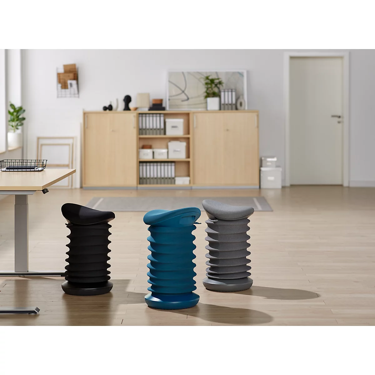 Three height-adjustable stools in black, blue and grey in front of an office background.