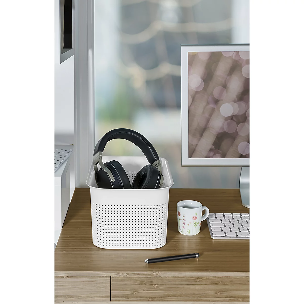 White basket with headphones on a wooden desk. Next to it, a mug and a pen. In the background, a computer monitor.