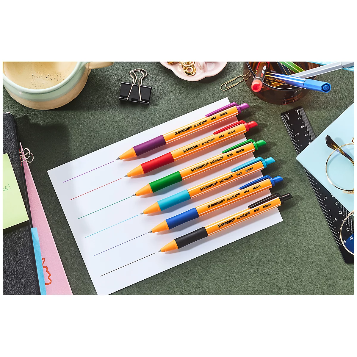 Des stylos multicolores sont sur du papier ligné, entourés de fournitures de bureau.