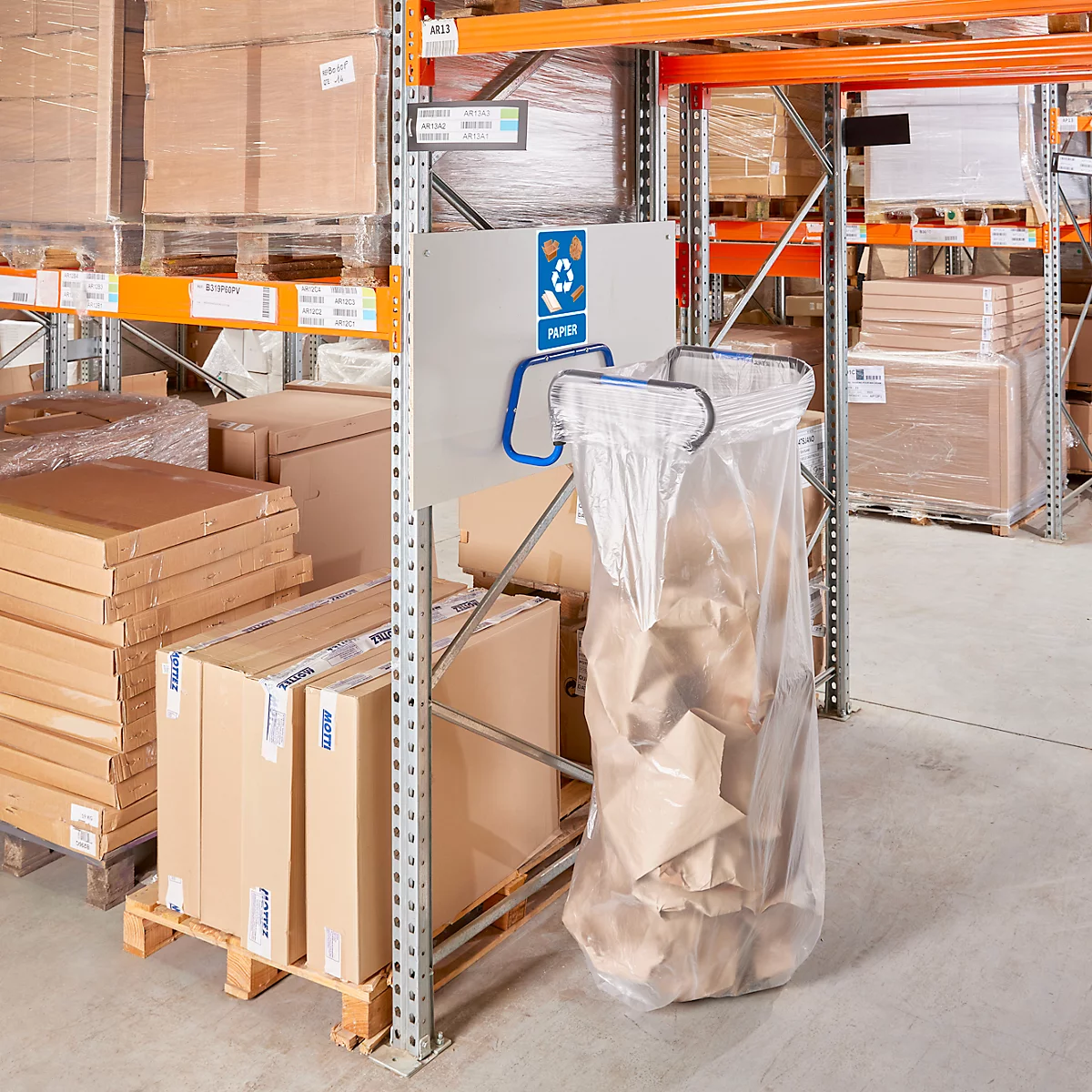 Poubelle à papier dans un entrepôt. Un sac à papier est suspendu à une structure métallique, à côté de panneaux. En arrière-plan, des étagères et des boîtes.