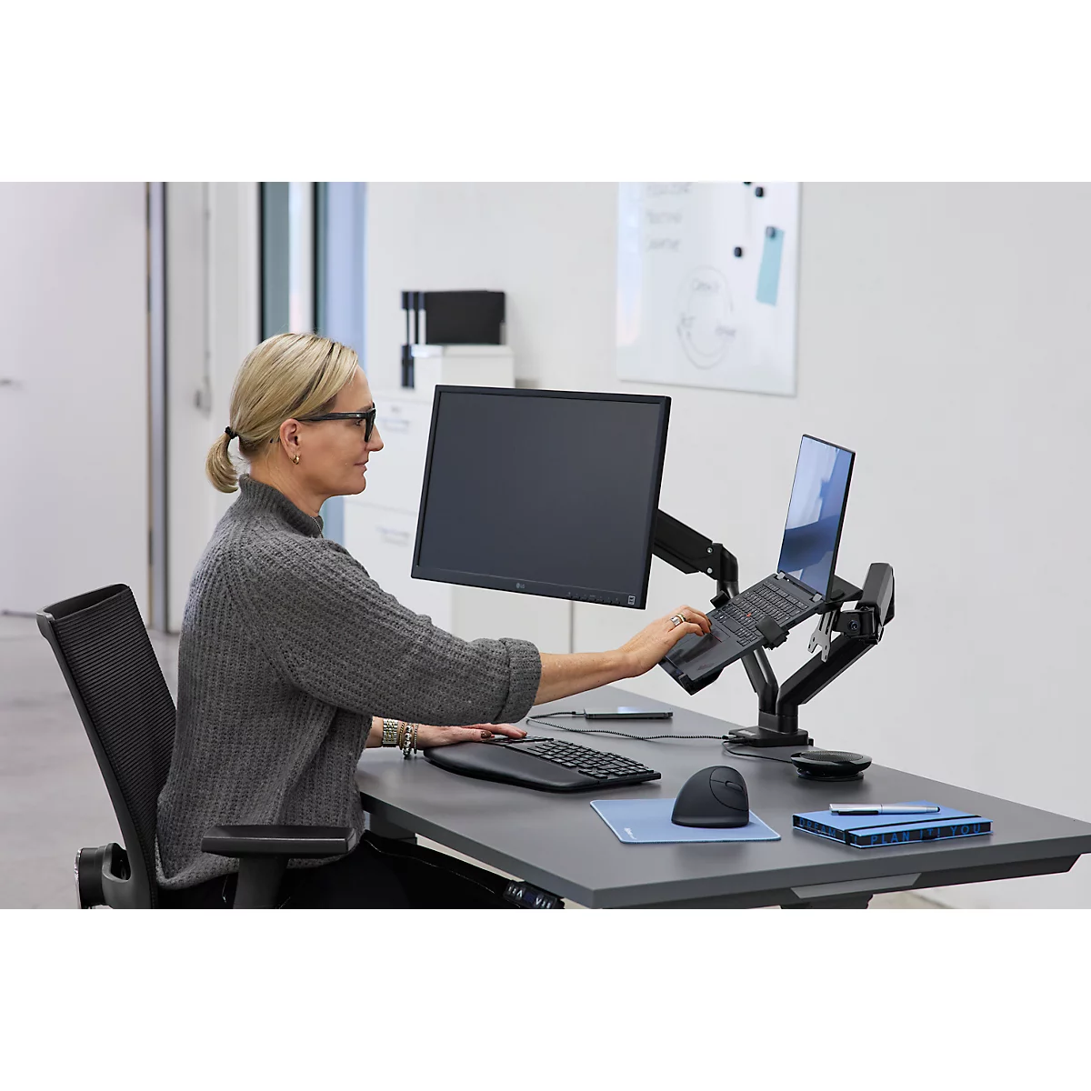 Femme travaillant à un bureau avec un ordinateur portable et un moniteur. Elle porte des lunettes et un pull gris.