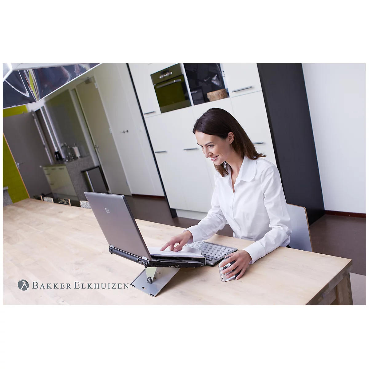 Une femme est assise à une table en train de travailler sur un ordinateur portable, souriante. Des accessoires sont sur la table. Une cuisine est visible en arrière-plan.