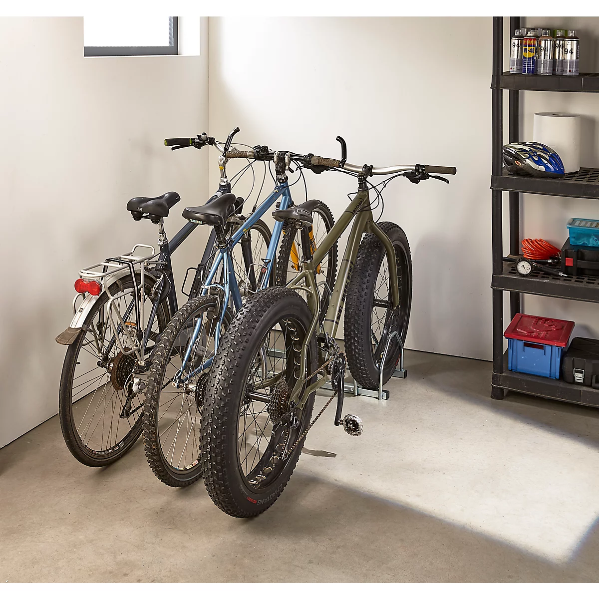 Trois vélos sont dans un garage. L'un est un fat bike, les autres sont des vélos normaux. A droite, une étagère avec des accessoires.