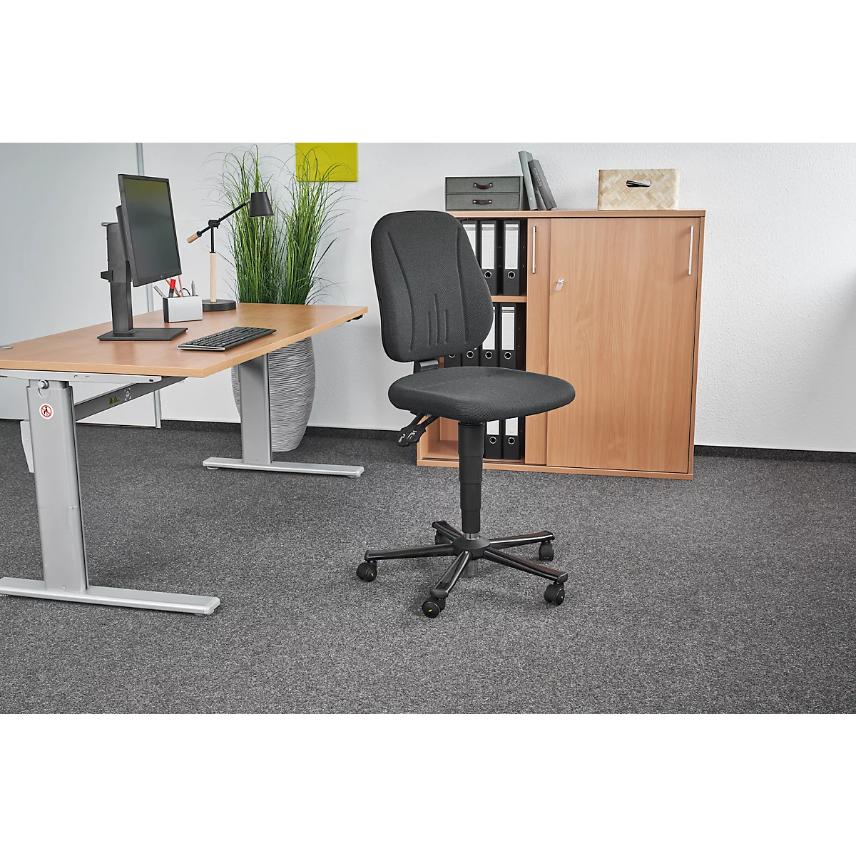 Office chair in an office, in front of a desk, cabinet and gray carpeted floor. Black chair with wheels, adjustable height, and padded seat and backrest.