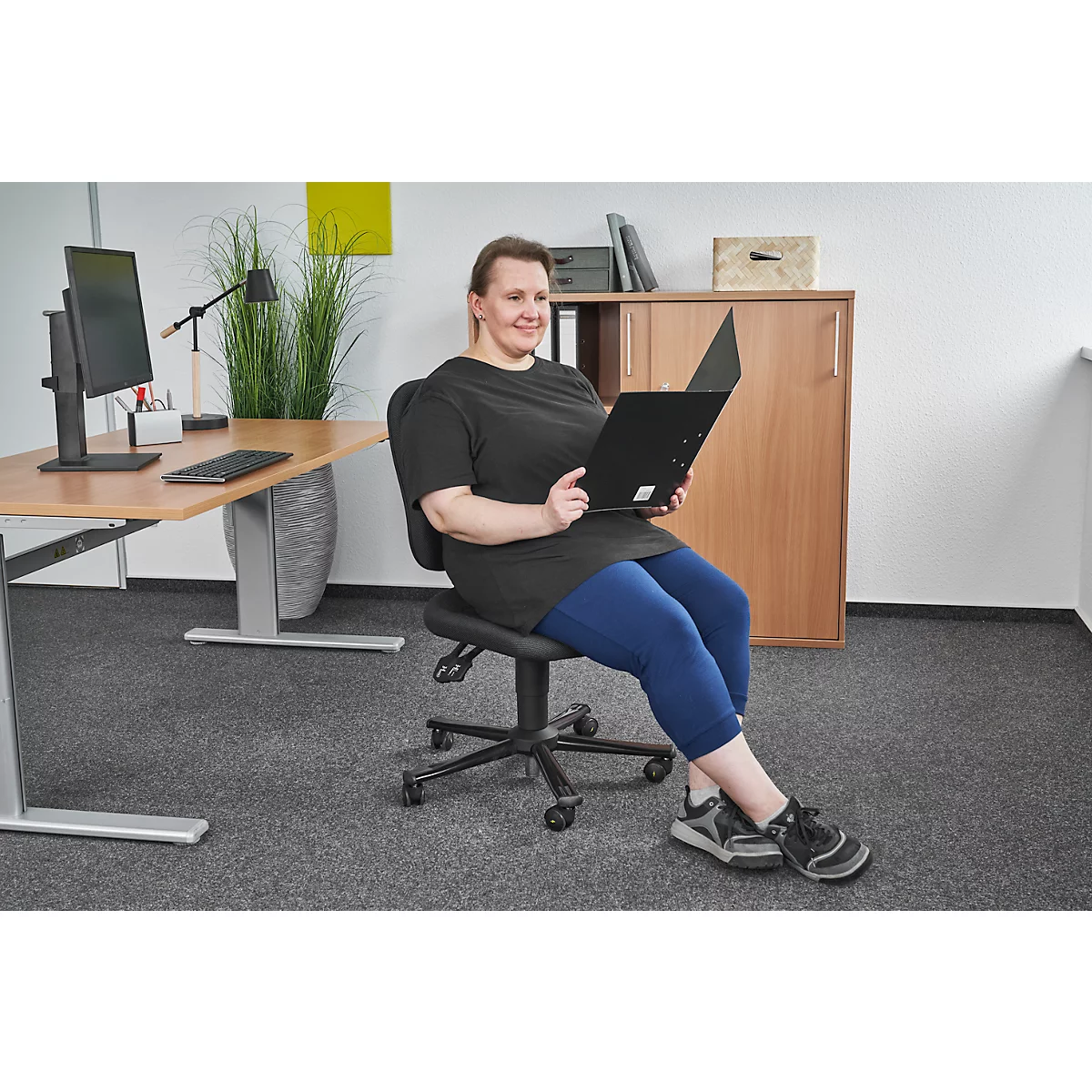 Woman sits on office chair, looks at file, smiling. Background desk, cabinet.