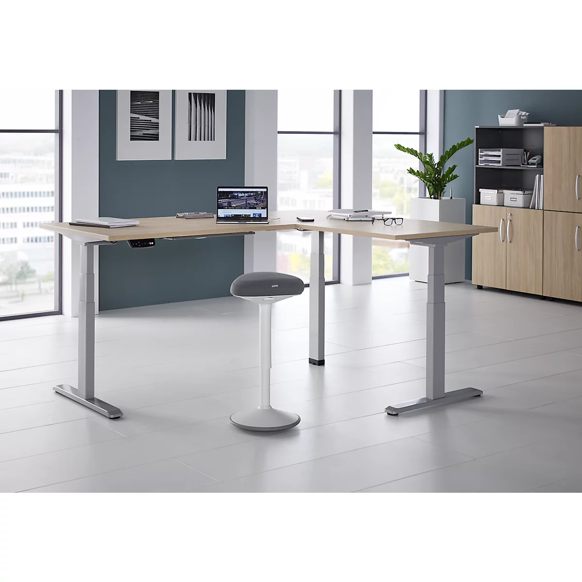 Un bureau d'angle réglable en hauteur avec un ordinateur portable, des lunettes et un tabouret dans un bureau moderne, environnement lumineux.