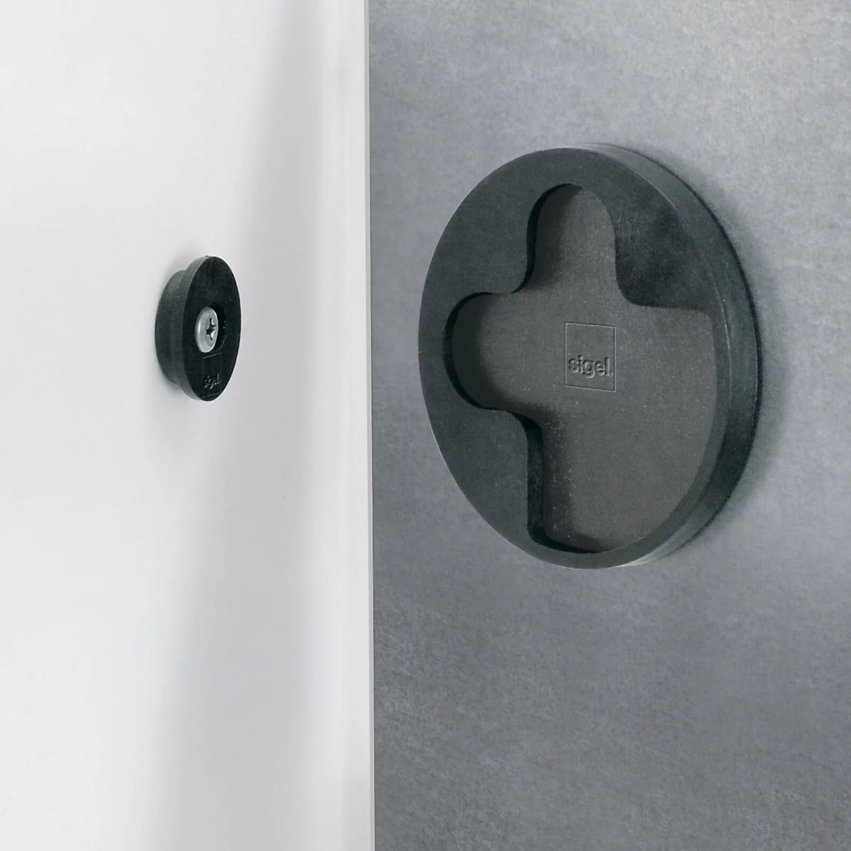 Deux objets noirs et ronds, le gauche petit et rond, le droit plat et en forme de croix. Ils sont fixés à un mur gris et à un objet blanc.