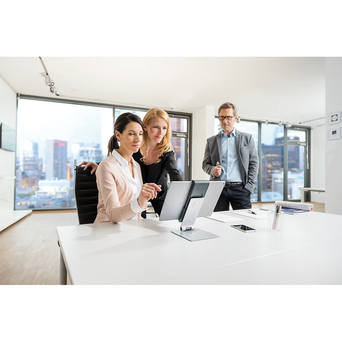 Three people looking at a tablet on a desk in the office. One woman is sitting, two are standing next to her.