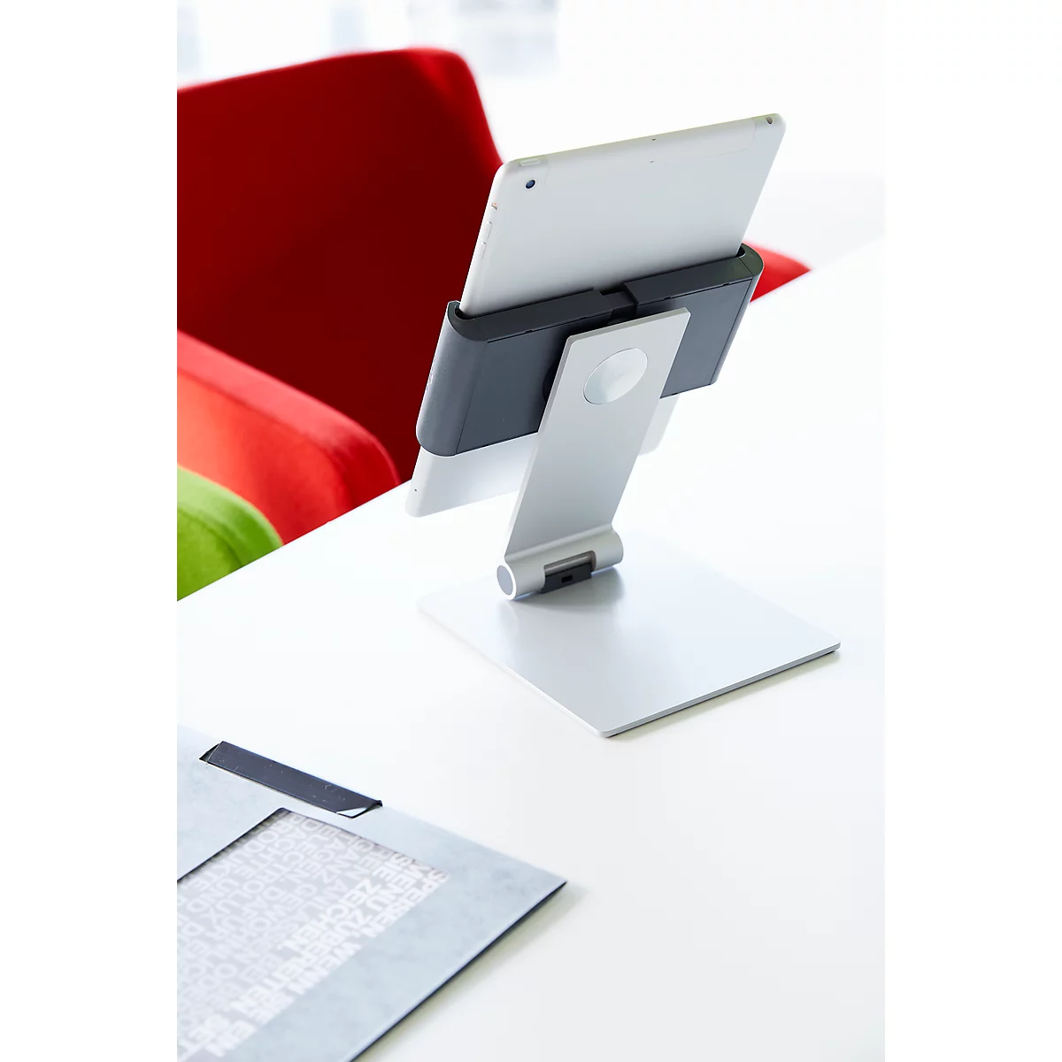 A tablet holder with tablet stands on a white table. In the background you can see a red armchair and a document.