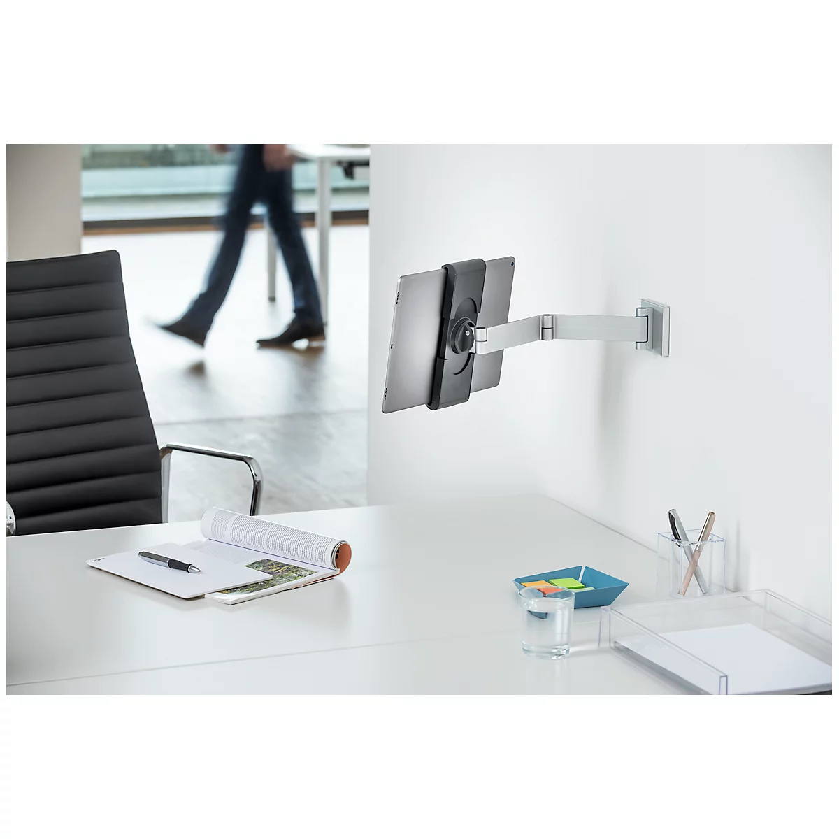 Tablet holder mounted on wall. Desk with notes, glass, pens. Person in background.