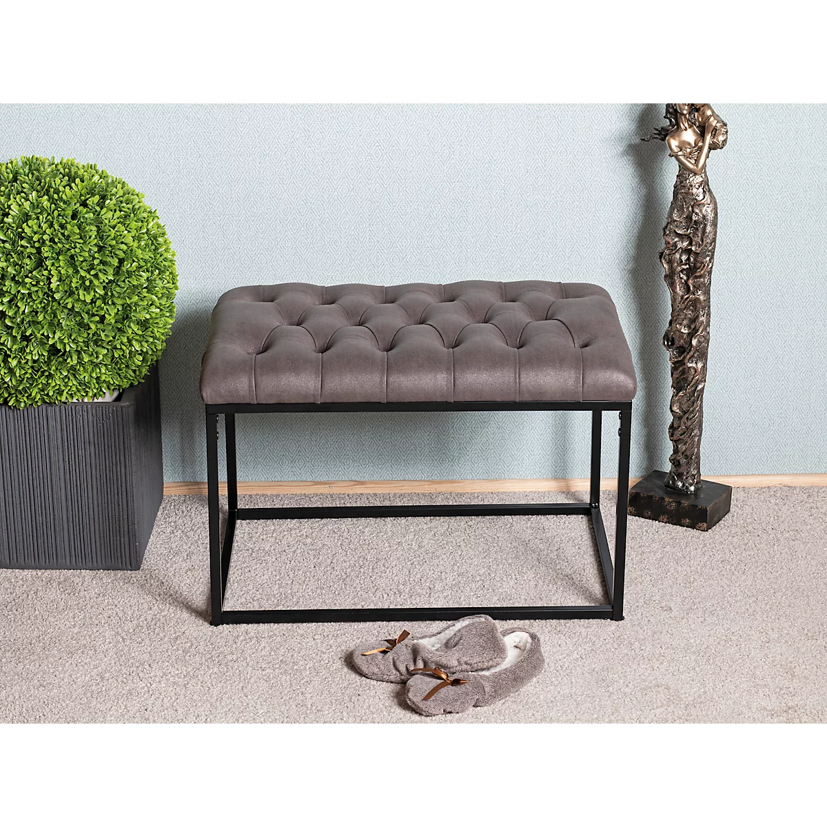 Banc rembourré gris avec cadre en métal noir. A côté d'un arbuste vert dans un pot sombre et d'une sculpture en bronze.