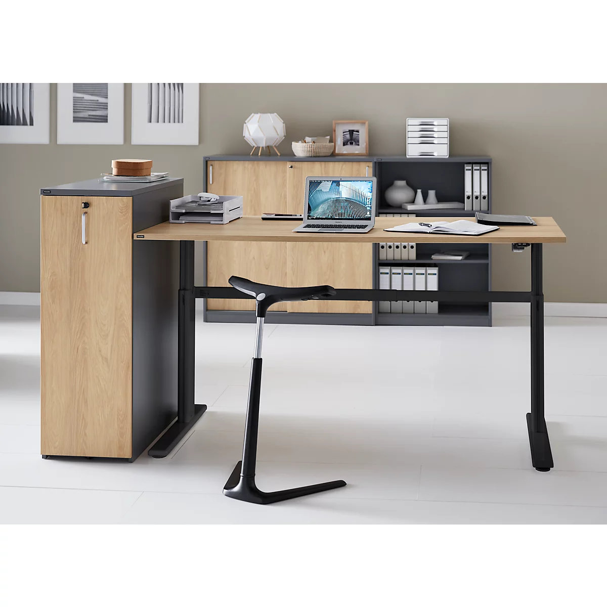 A modern desk with a laptop and standing aid in a bright office. The desk has a wood look, the frame is black.