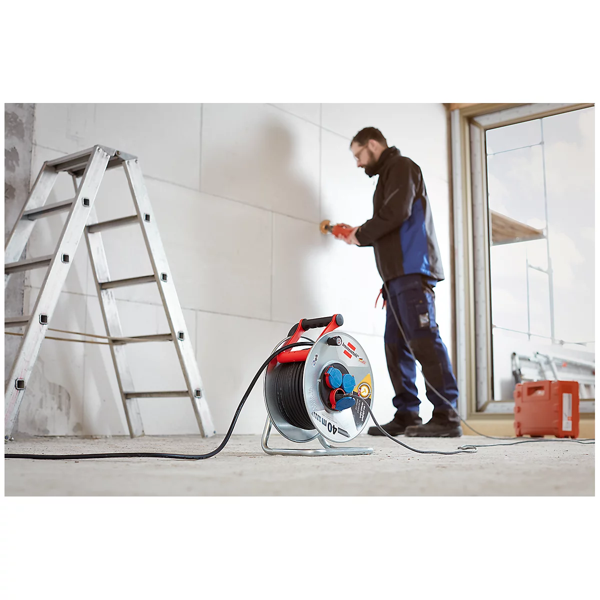 Un homme travaille sur un mur avec une ponceuse. Une bobine de câble et une échelle sont dans l'image.