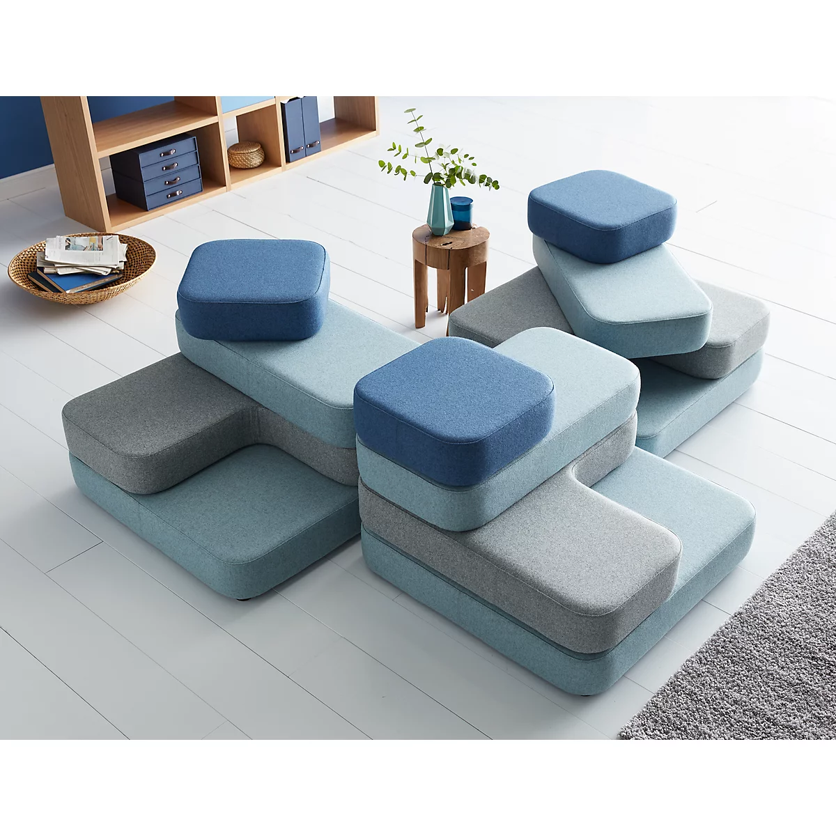 Modular sofa system in blue and grey. Multiple elements form a seating group on a light floor. Shelf and decoration in the background.