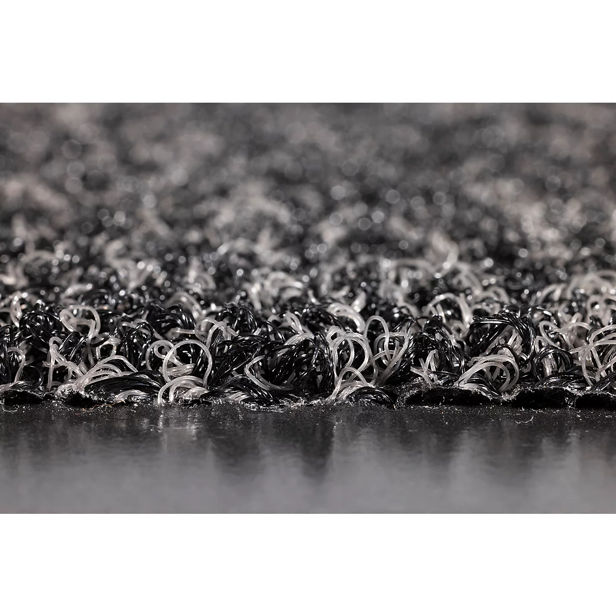 Gros plan d'un paillasson à la texture composée de boucles en plastique noir et gris entrelacées.
