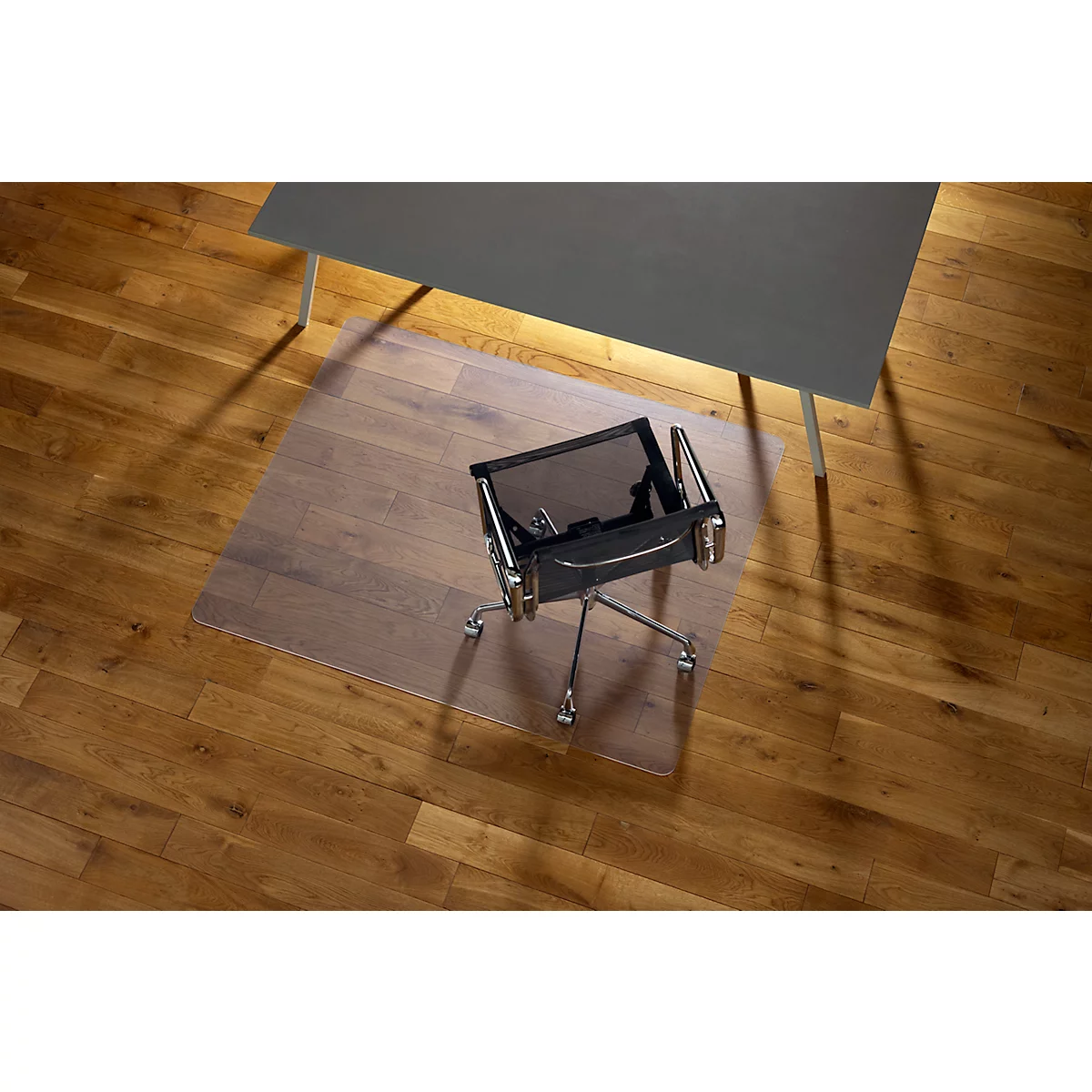 Chaise de bureau sur un tapis de protection de sol transparent sous un bureau gris. L'arrière-plan est un plancher en bois.