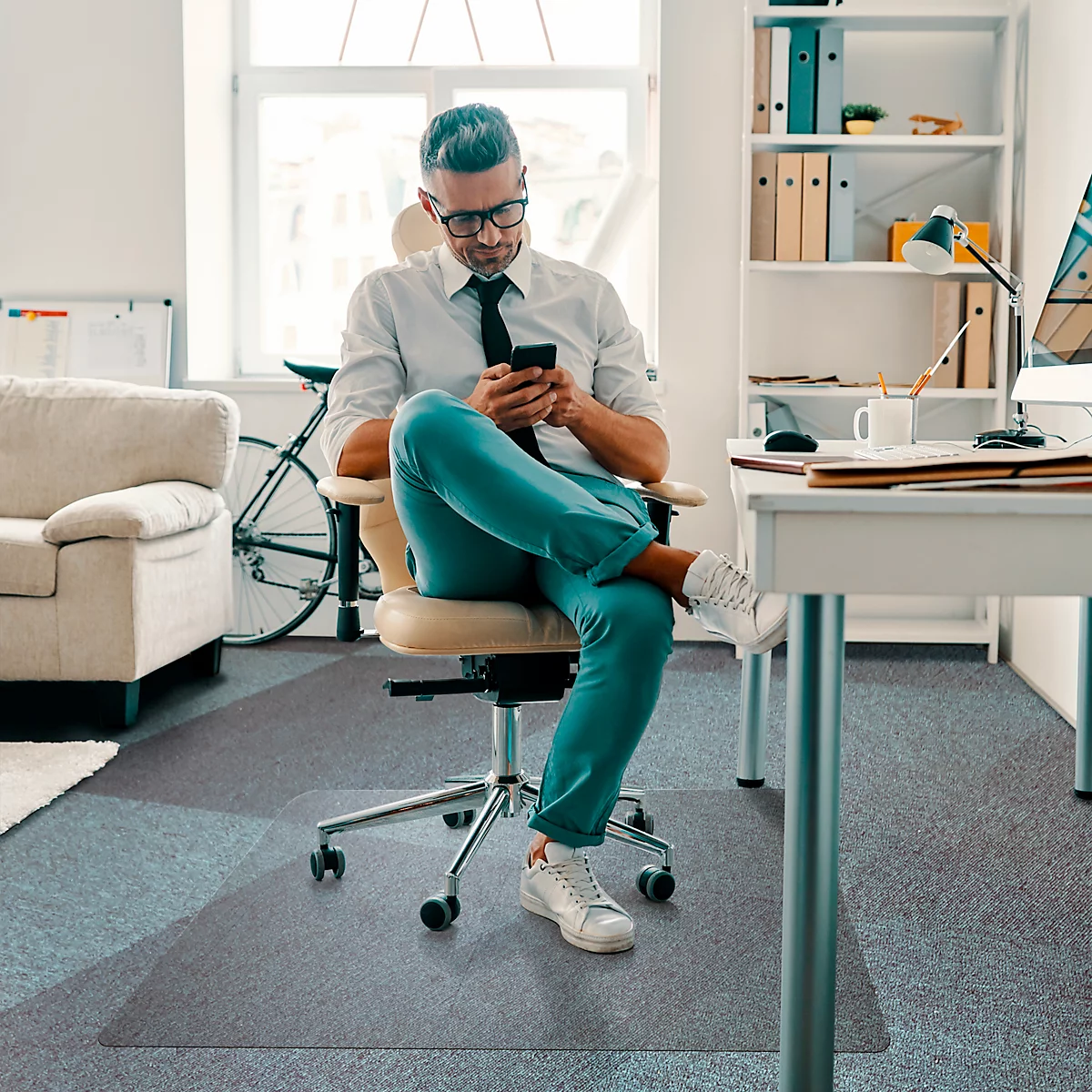 Un homme en chemise est assis sur une chaise de bureau et regarde son téléphone. Pantalon clair, lunettes et baskets.