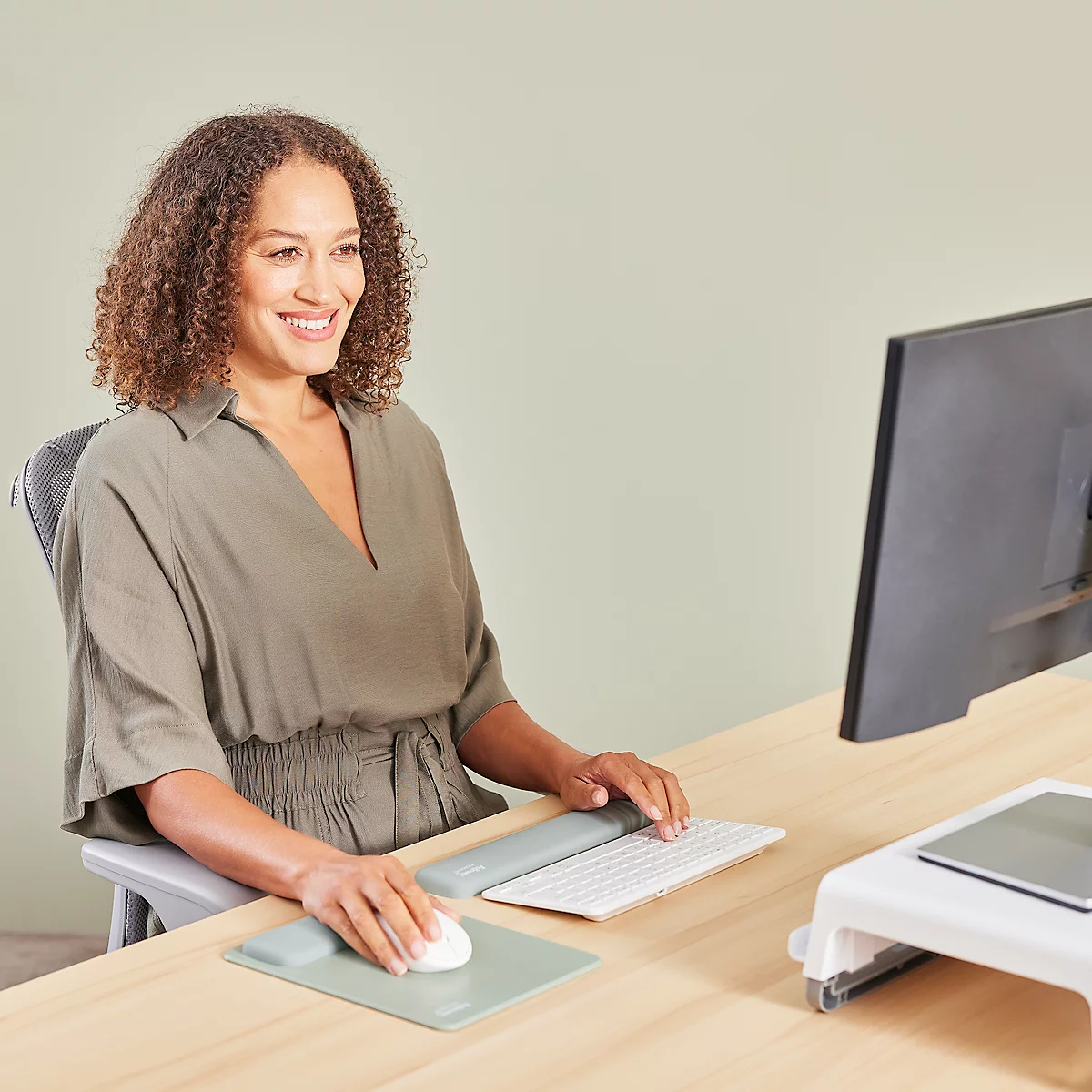 Une femme souriante est assise à un bureau, travaillant sur un ordinateur. Elle porte un haut vert et a les cheveux bouclés. Il y a une souris, un clavier et un écran sur le bureau.