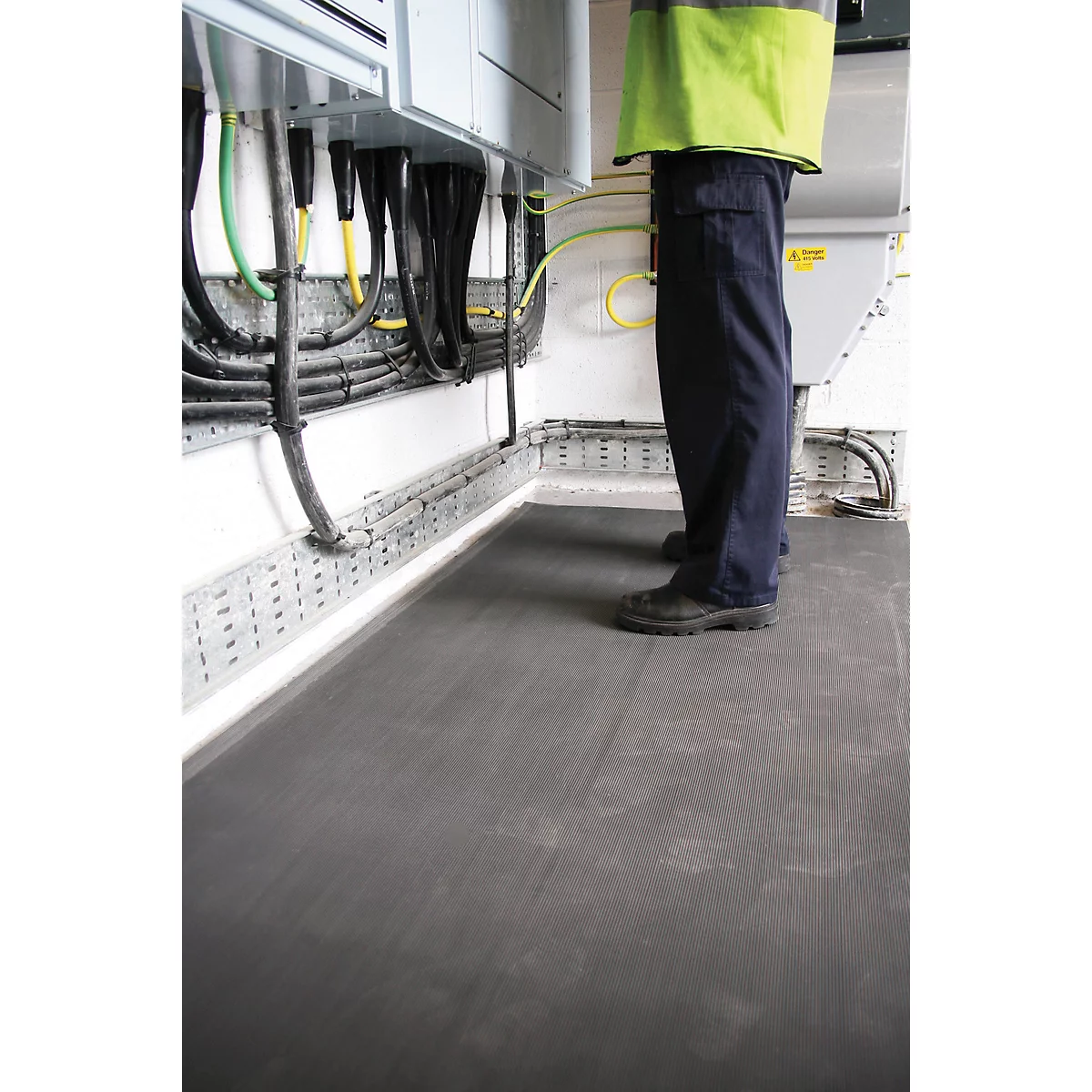 Un travailleur se tient sur un tapis noir près de câbles et d'équipements électriques. Il porte des vêtements sombres et un gilet jaune.