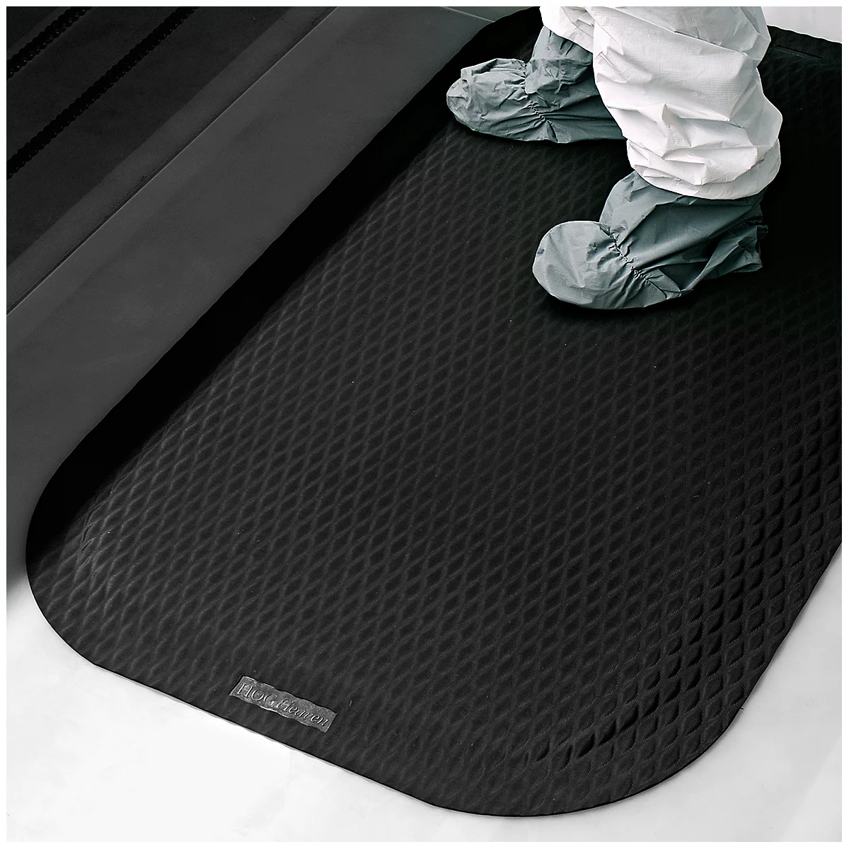 Une personne en combinaison blanche et couvre-chaussures se tient sur un tapis noir et strié. Un élément noir est visible en arrière-plan.