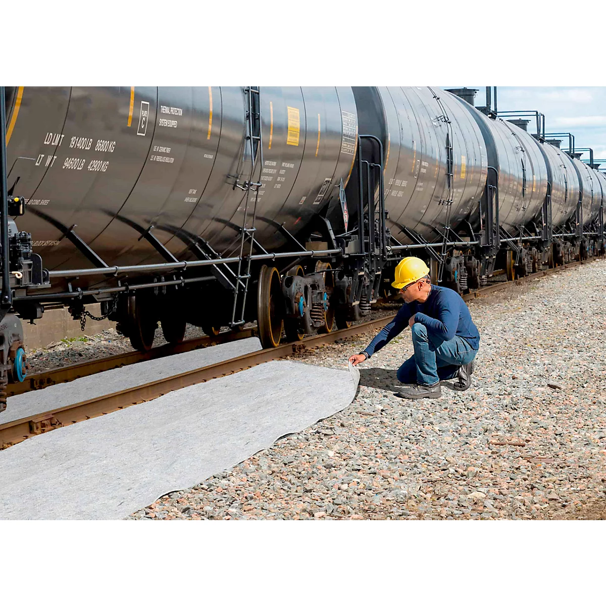 Un homme portant un casque jaune est agenouillé à côté d'une voie ferrée. Il examine un tissu gris qui se trouve entre les rails.