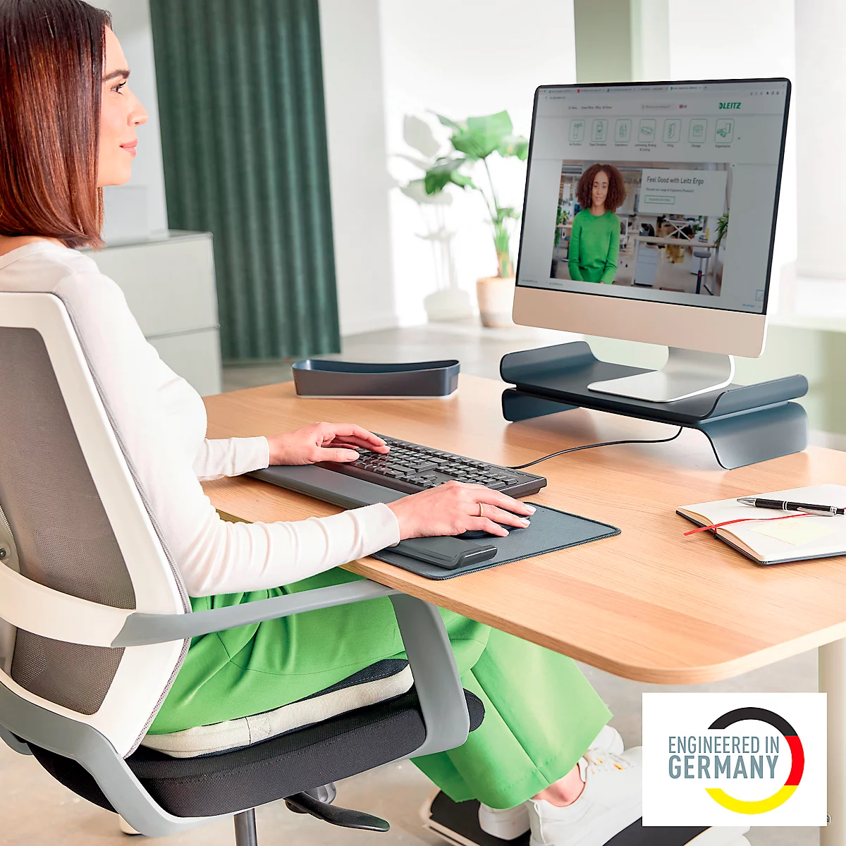 Une femme travaille sur un ordinateur posé sur un support. Elle porte un chemisier blanc et un pantalon vert.