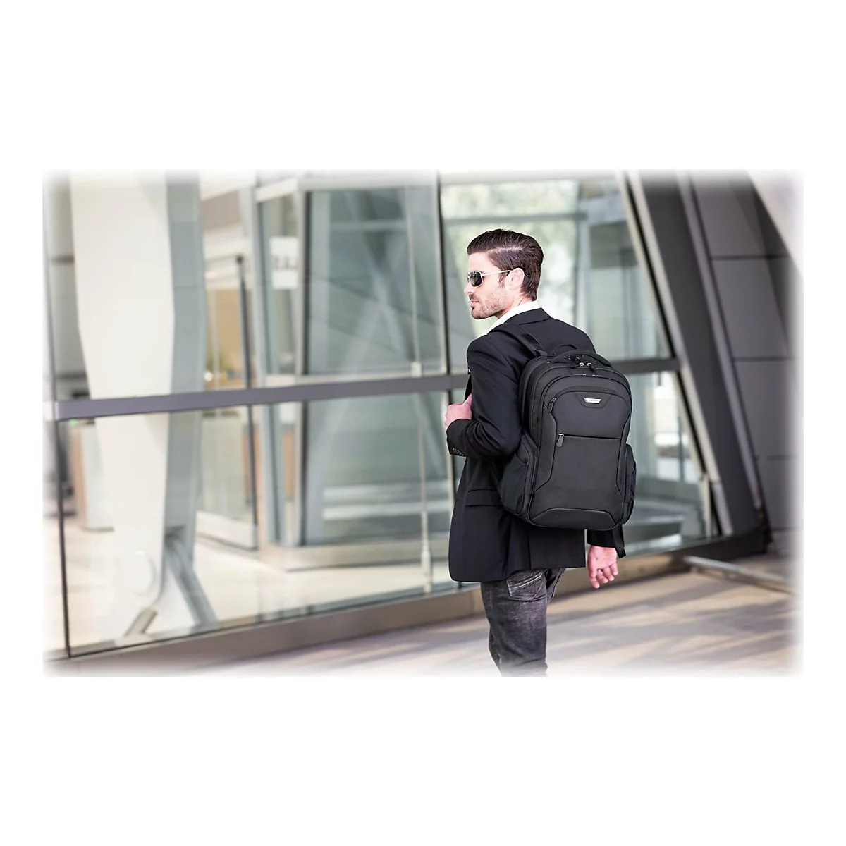 Mann mit Rucksack, Sonnenbrille und Anzug, der an einem sonnigen Tag vor einem modernen GebÃ¤ude steht.