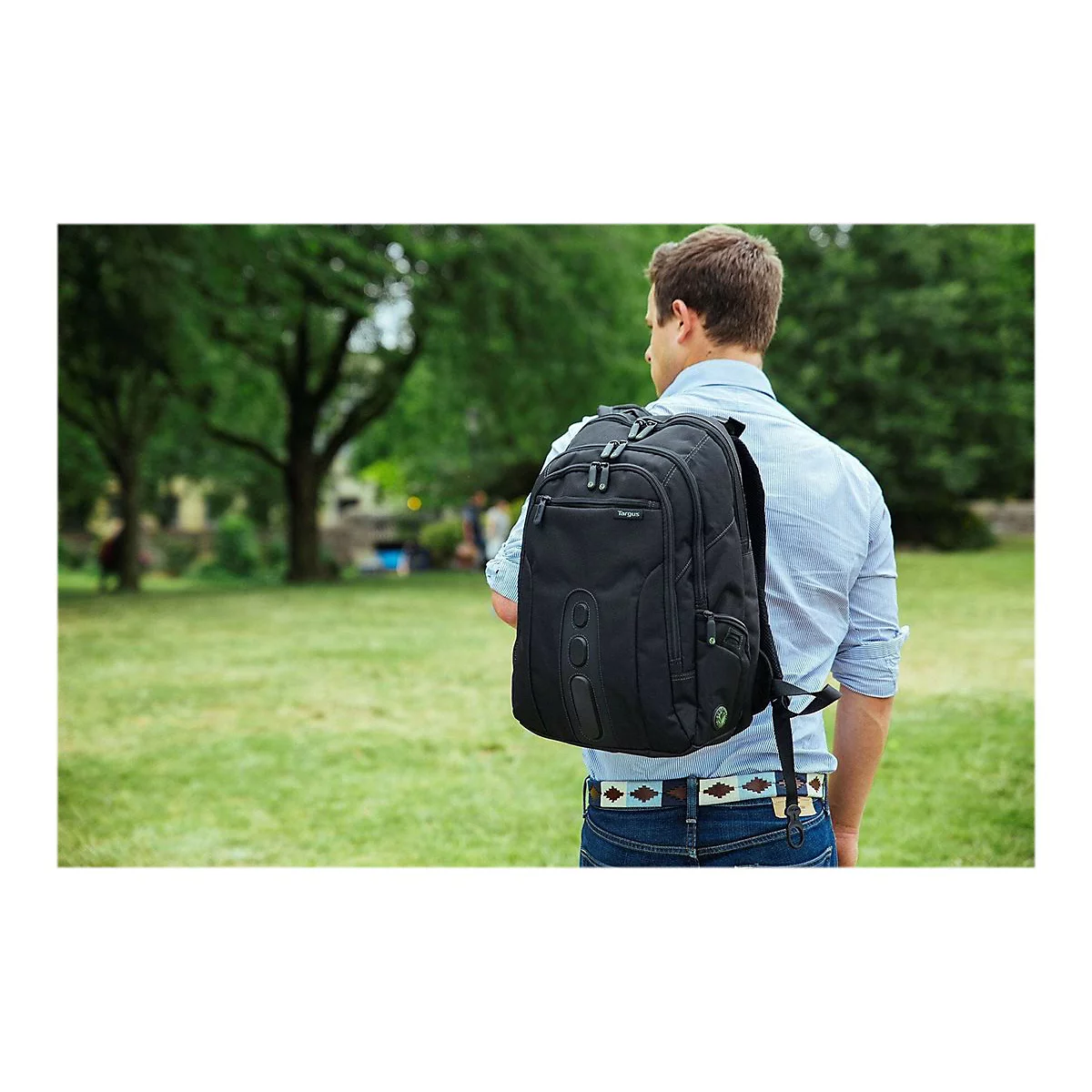 Ein Mann, der einen schwarzen Rucksack auf dem Rücken trägt, geht durch einen Park. Der Rucksack hat das Logo 'Targus'.