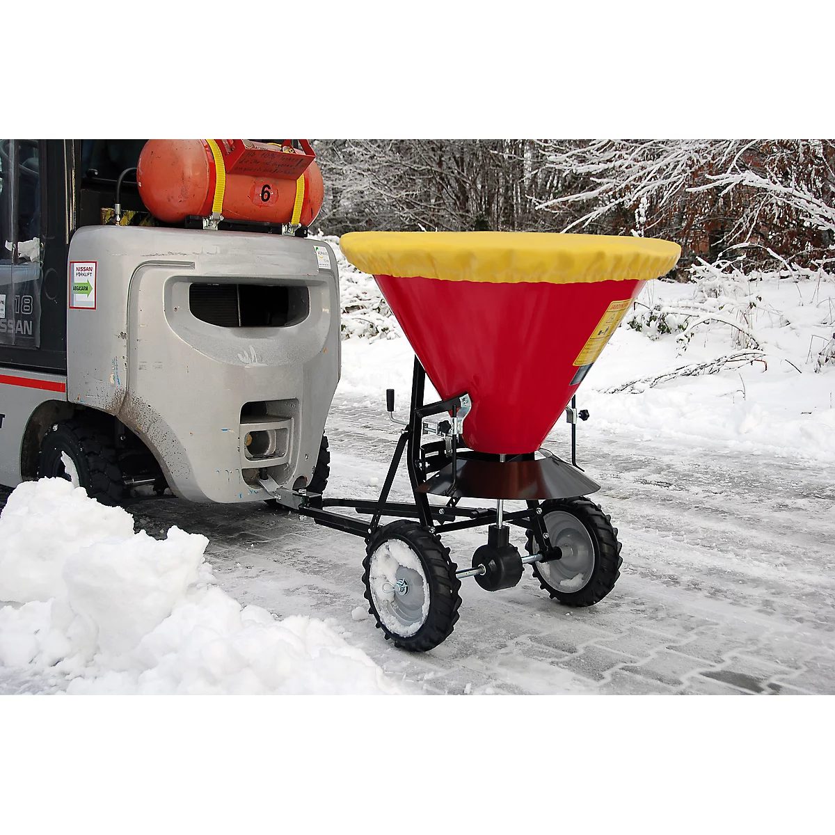 Forklift pulling a trailer with a salt spreader. On the trailer a red container with a yellow lid. Snow covers the ground.