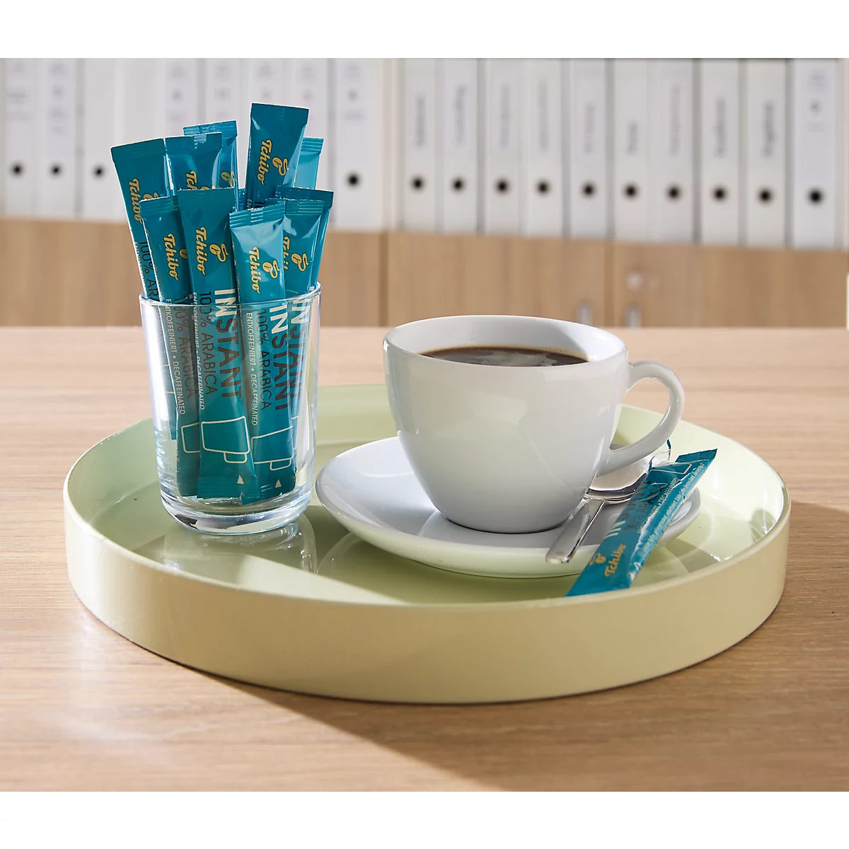 Ein hellgelbes Tablett steht auf einem Holztisch mit einer Tasse Kaffee und einem Glas voller blauer Kaffeesticks.