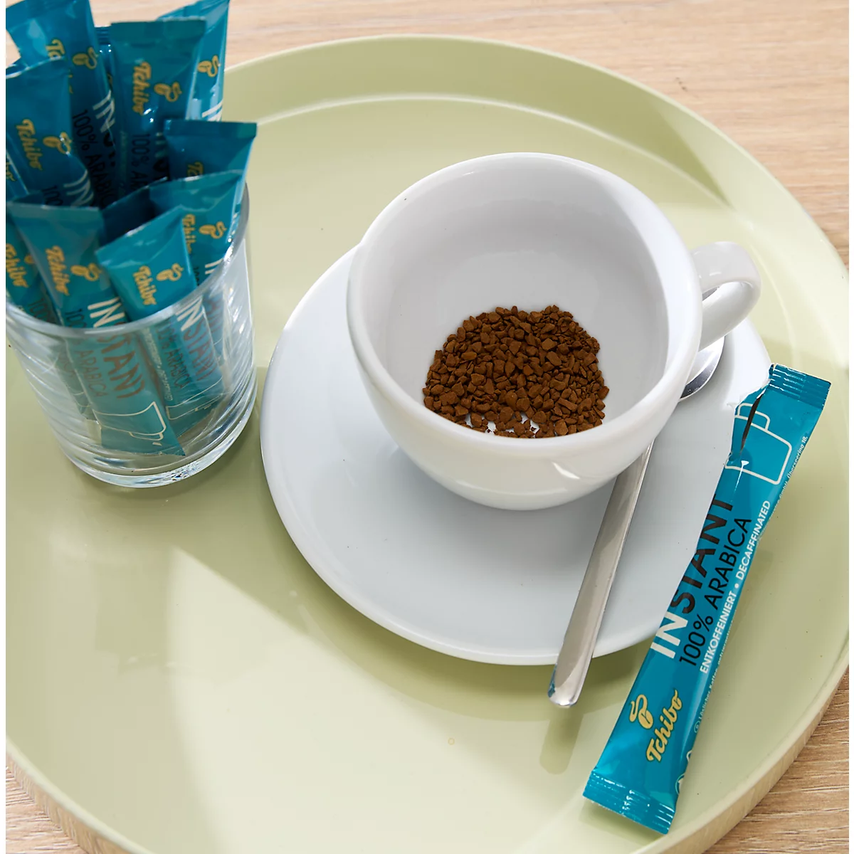 Weiße Kaffeetasse mit Instantkaffeepulver auf einem hellgrünen Tablett, daneben Tchibo Kaffeesticks im Glas.