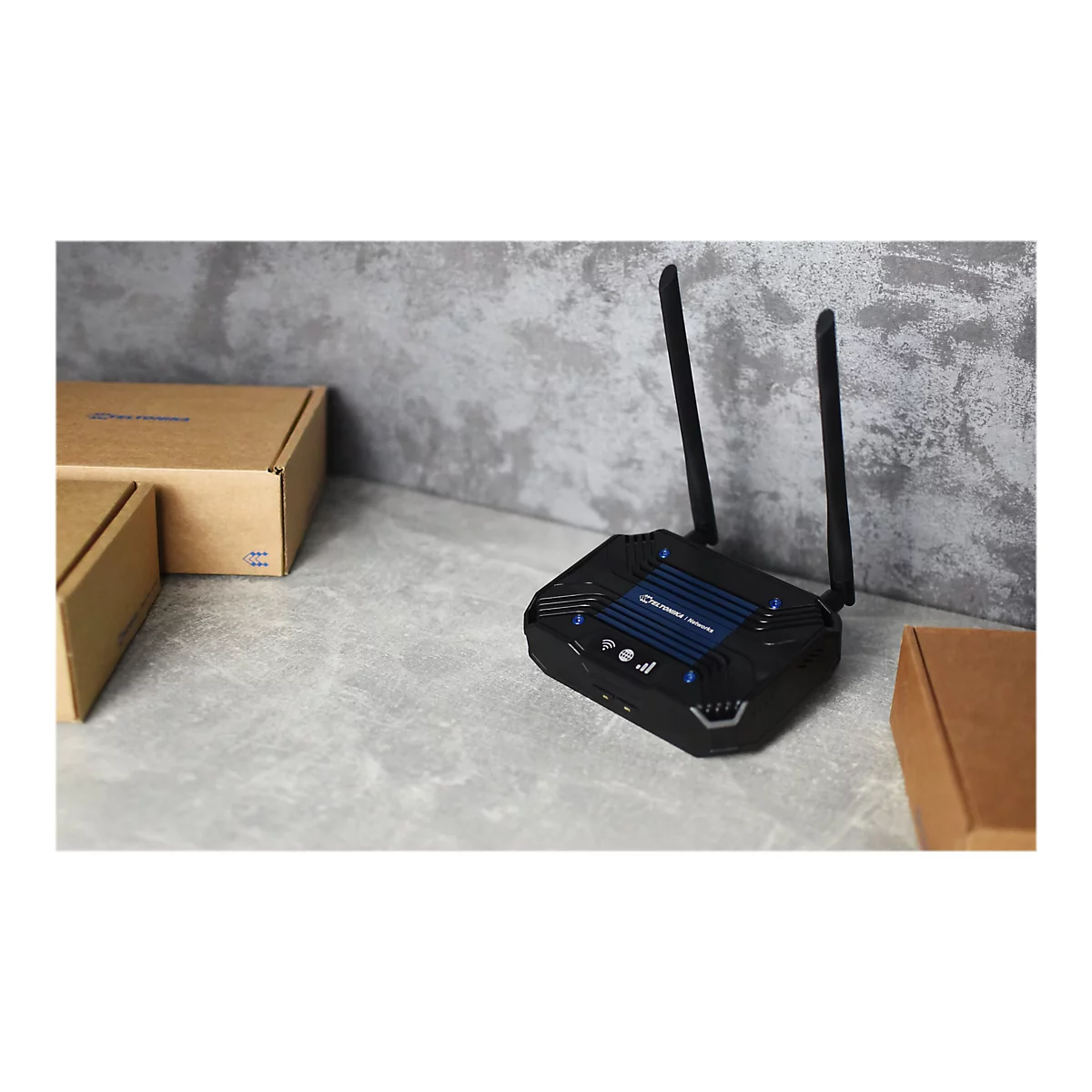 Schwarzer Router mit zwei Antennen. Daneben sind zwei braune Kartons zu sehen. Der Router steht auf einer grauen OberflÃ¤che.