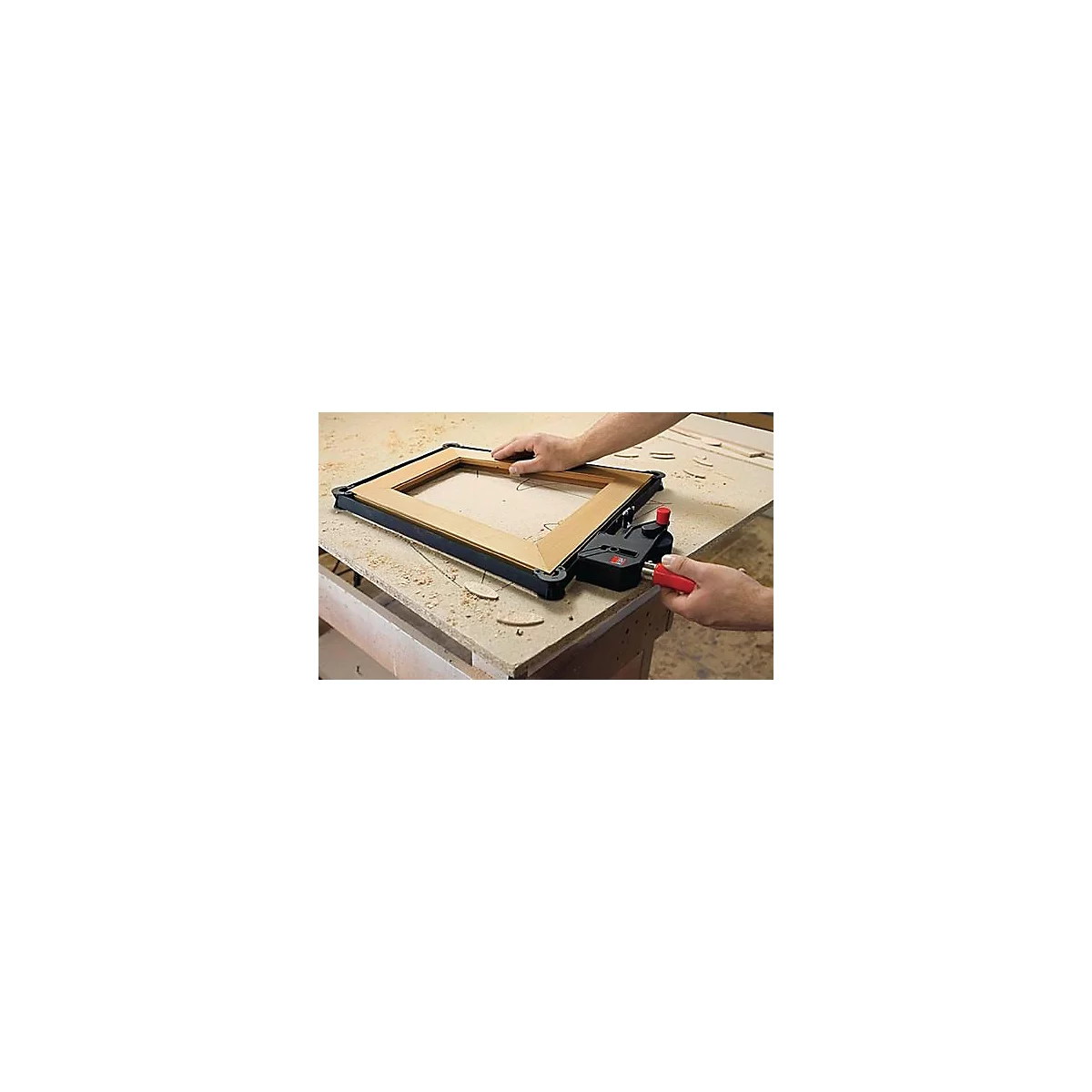 Des mains travaillent sur un cadre en bois avec un connecteur d'angle noir et un outil rouge sur un établi. Des morceaux de bois sont posés à côté.