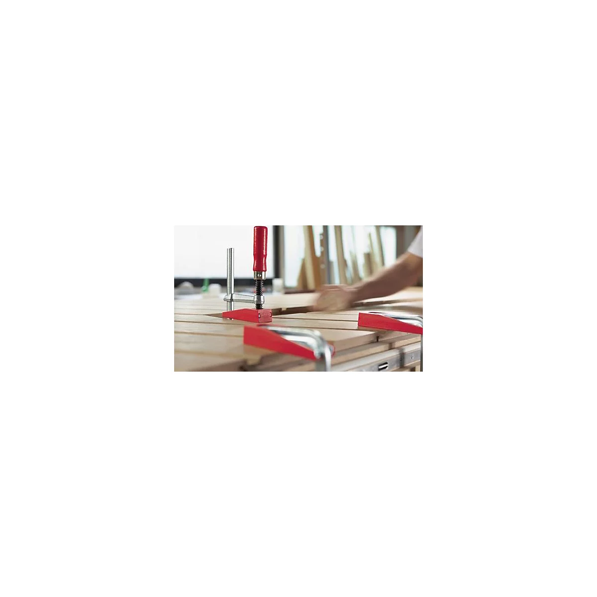 Gros plan sur des serre-joints rouges sur une surface en bois. On voit un homme travailler en arrière-plan.