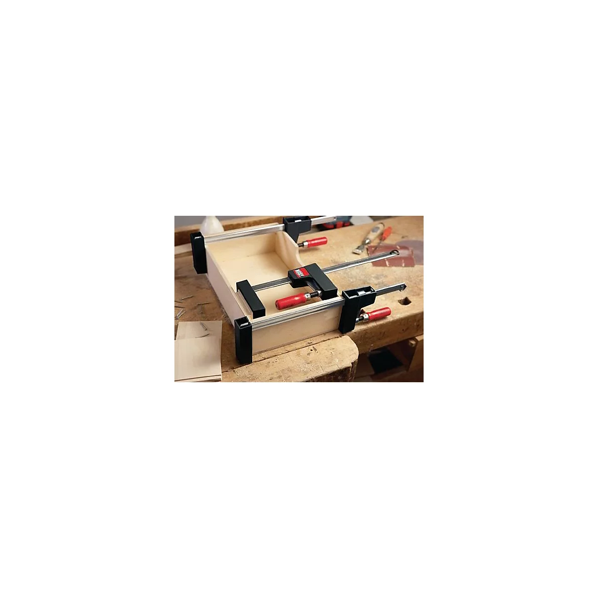 Une boîte en bois est collée avec des pinces. Poignées rouges, supports noirs sur une surface en bois clair.