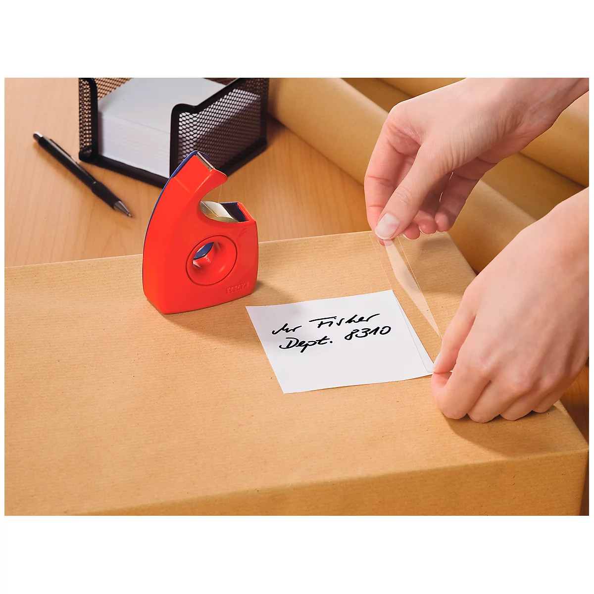 Hands are taping a sticker with text to a cardboard box. Next to it tape dispenser and notepad.