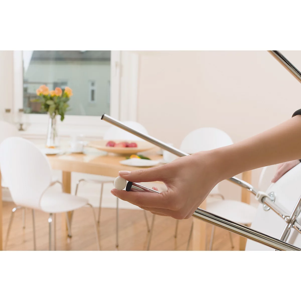 Eine Person hält ein Stuhlbein-Detail. Im Hintergrund: Tisch mit Essen, Stühle, Fenster.