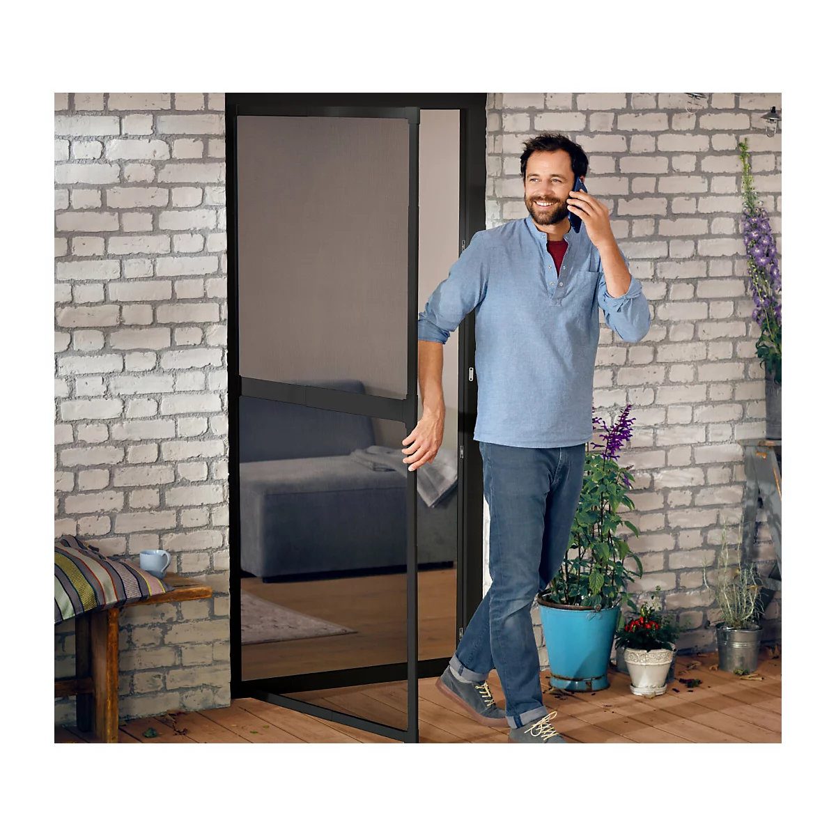 Ein Mann telefoniert in einem Türrahmen. Er steht auf einer Holzterrasse vor einer Backsteinwand.