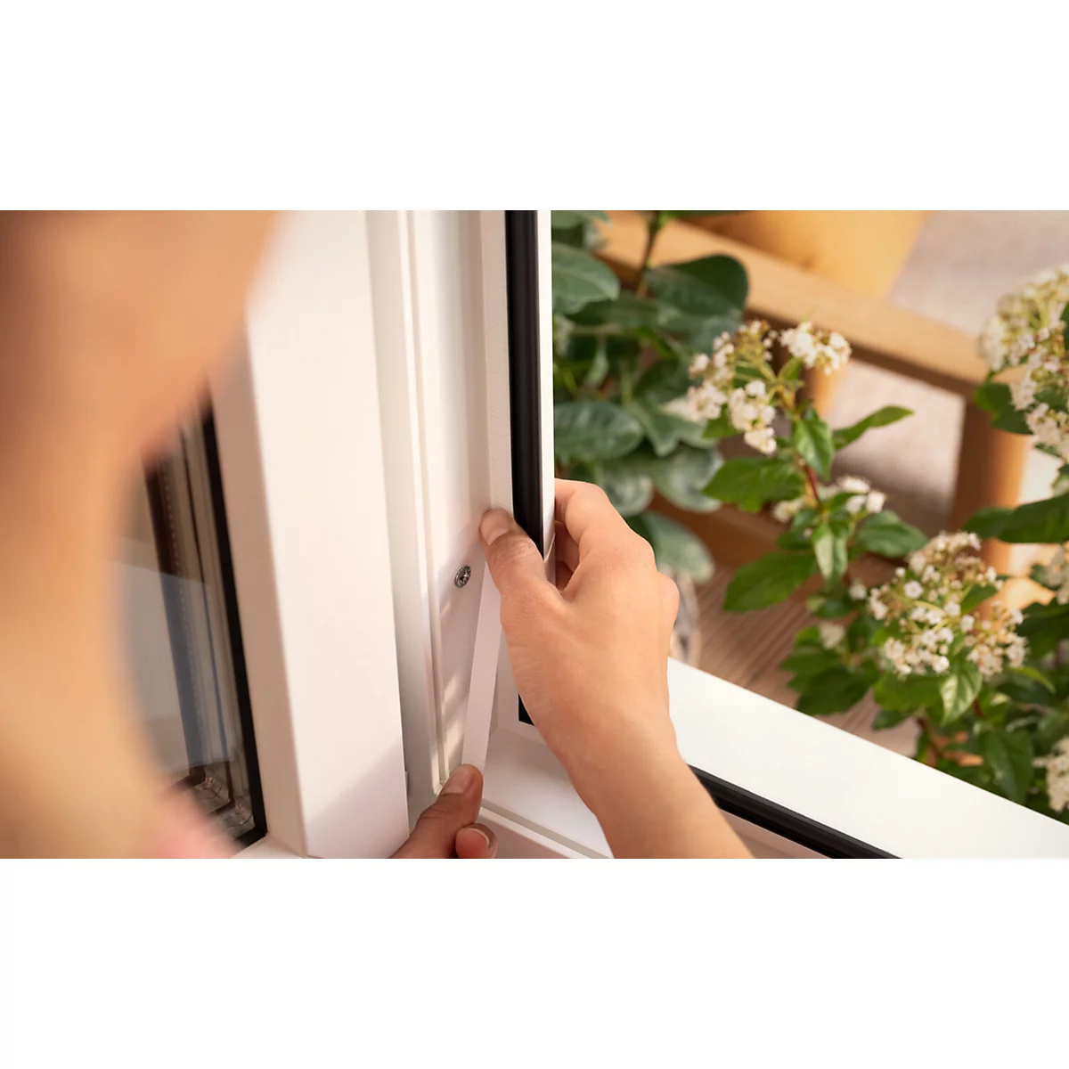 Nahaufnahme einer Person, die eine Dichtung in einem weißen Fensterrahmen anbringt. Im Hintergrund grüne Pflanzen und ein Holztisch.