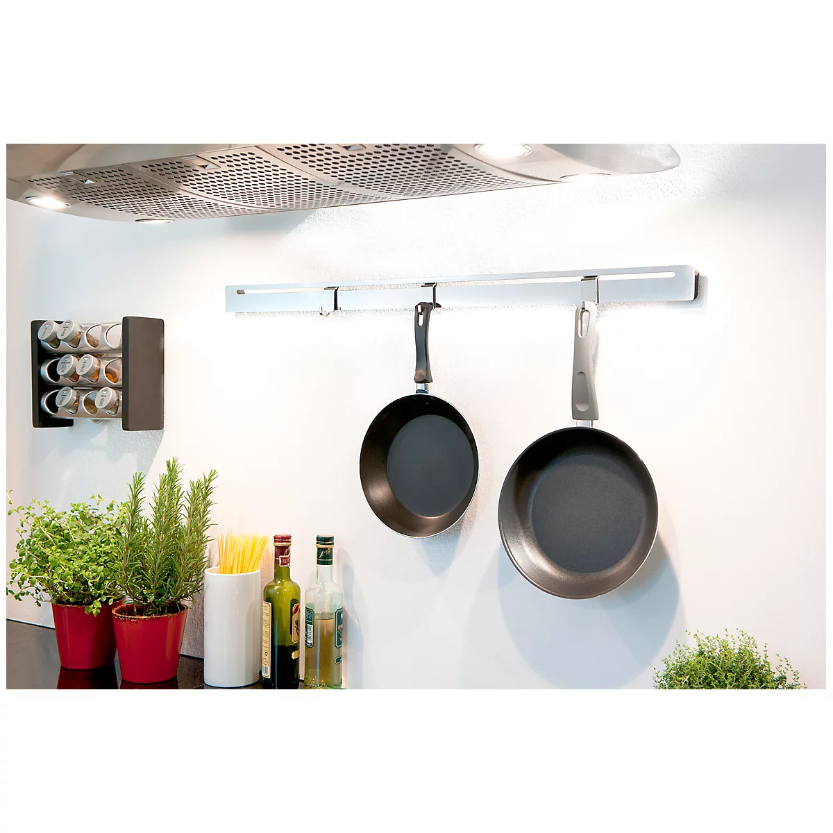 Two pans hang on a rail in a kitchen. Below are spices, herbs and oil bottles.